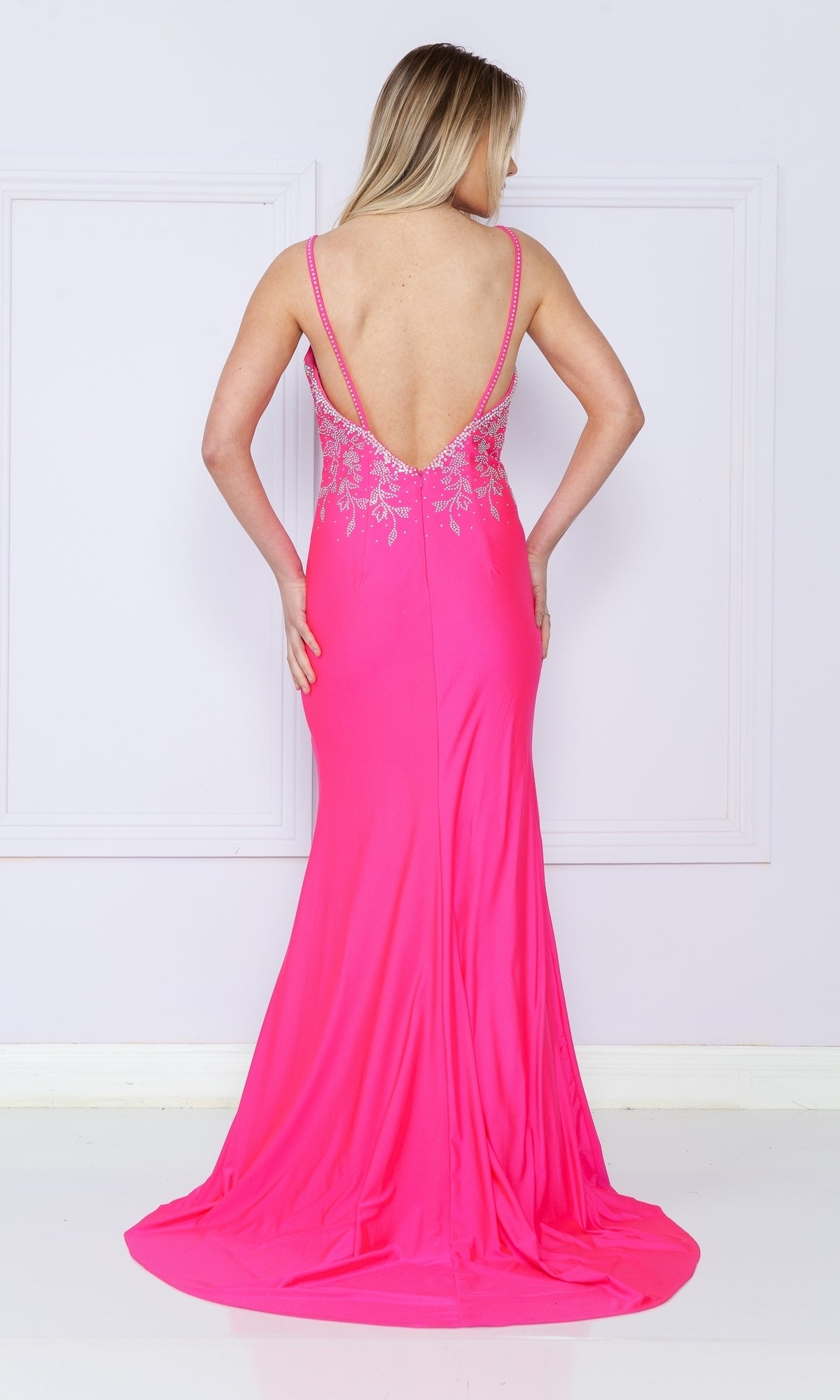 A woman stands against a white wall in the Backless Long Jersey Prom Dress: Poly USA 9120, a bright pink, floor-length gown with an open back, floral embellishments, and a subtle thigh-high slit.